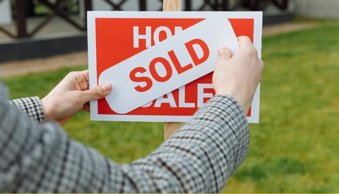 Person holding a real estate sold sign in front of a house with green lawn