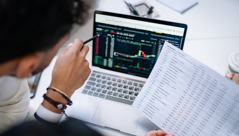Person viewing stock market charts on laptop while holding financial data sheet with graphs and numbers